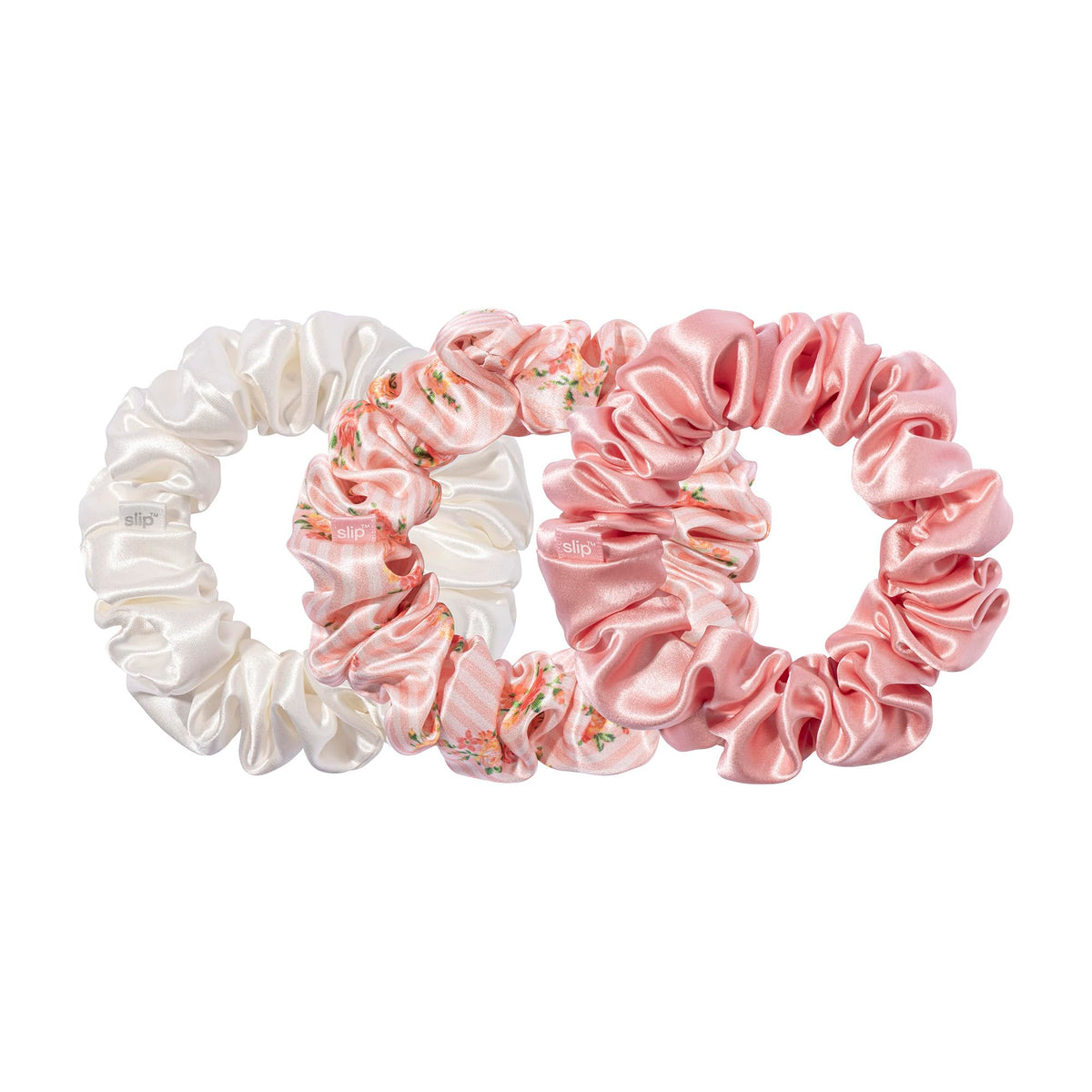 Three large gathered scrunchies in ivory, pink floral print, and solid pink, with small slip labels, overlapping on a white background