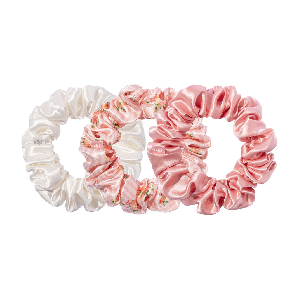 Three large gathered scrunchies in ivory, pink floral print, and solid pink, with small slip labels, overlapping on a white background