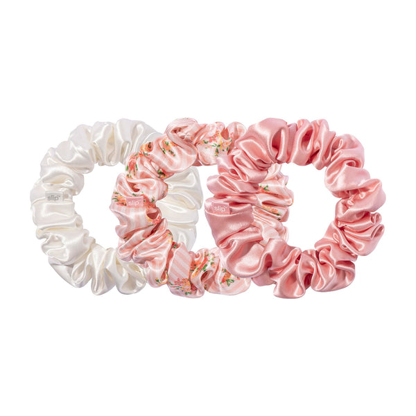 Three large gathered scrunchies in ivory, pink floral print, and solid pink, with small slip labels, overlapping on a white background