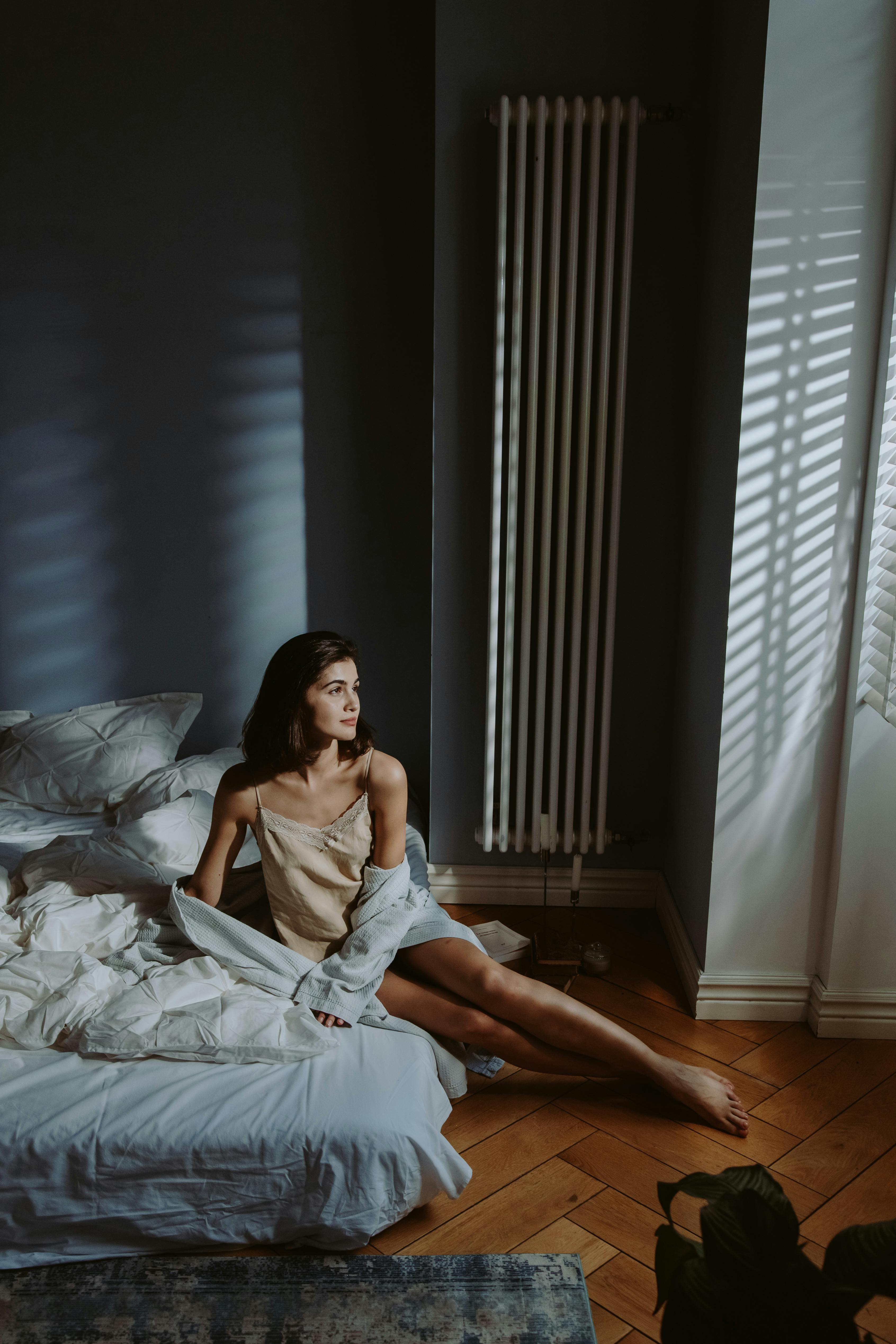 Woman sitting on bed in softly lit room with window blinds casting shadows on the wall and floor