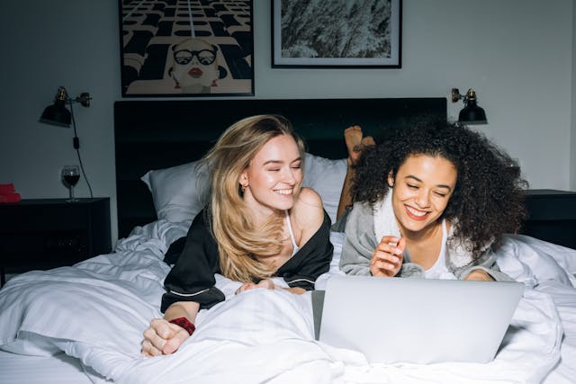Two women lying on a bed together, smiling and looking at a laptop screen in a cozy bedroom setting.