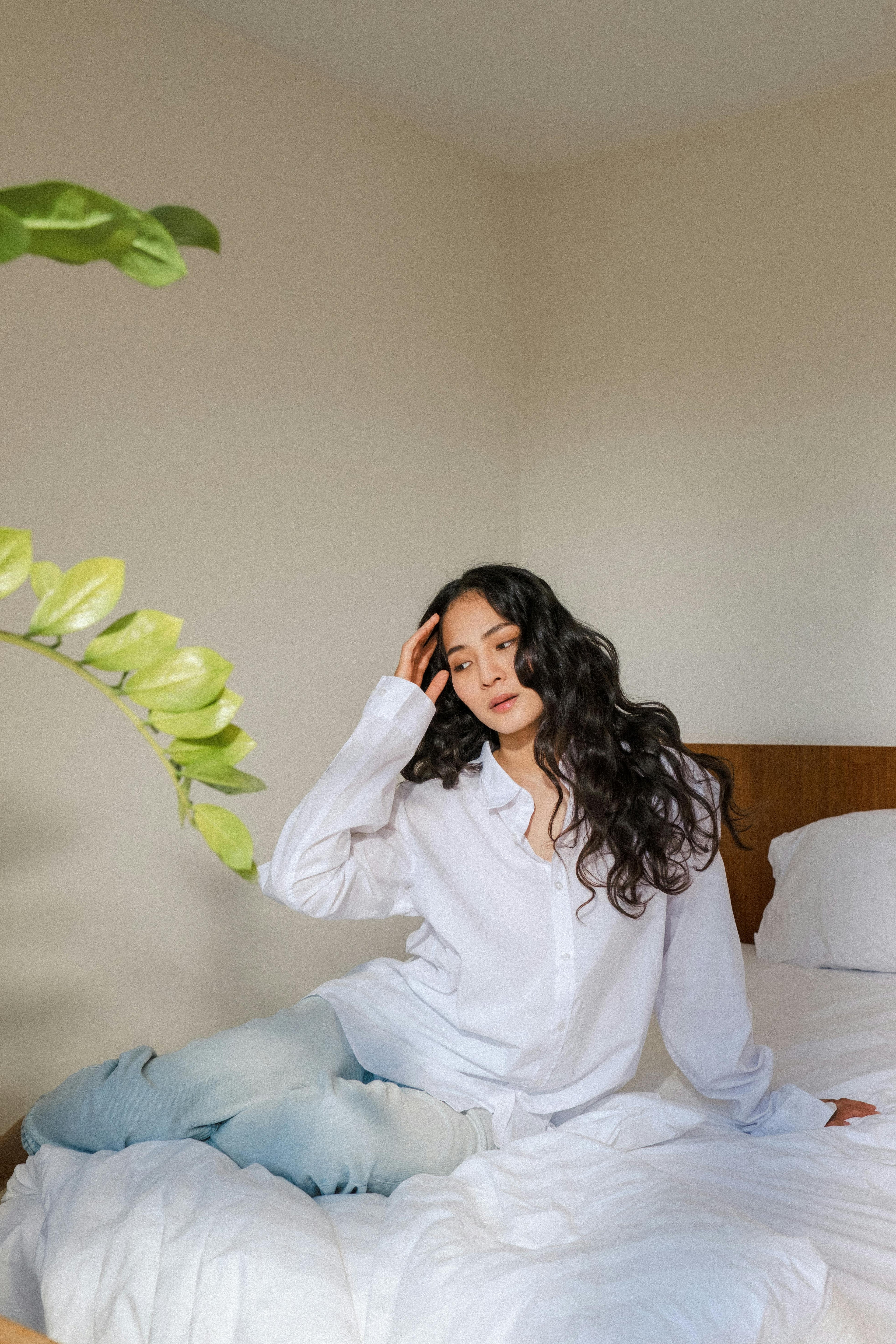 Woman with long wavy hair in a white shirt sitting thoughtfully on a bed with white bedding near a leafy plant
