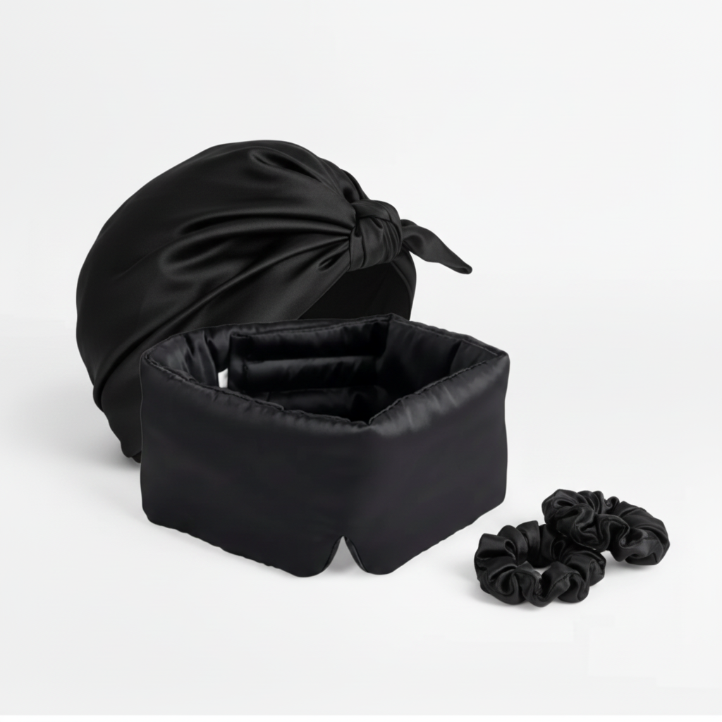 Black silk sleep bonnet, padded eye mask, and two silk scrunchies arranged on white background